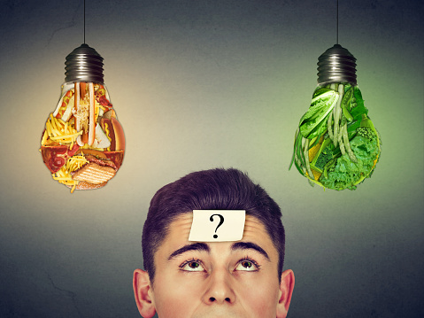 man with question mark on forehead thinking looking up at junk food and green vegetables light bulbs isolated on gray background. Diet choice right nutrition concept