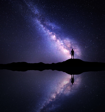 Night purple sky with Milky Way, stars and silhouette of a standing man on the mountain near the lake with sky reflection in water. Milky way and man on the hill. Galaxy and man. Tourism. Space