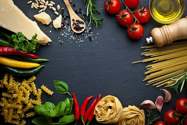 Ingredients for cooking traditional italian pasta shot on dark stone background. The ingredient are placed all around the frame leaving a useful copy space at the center. Composition includes tagliatelle pasta, spaghetti, olive oil, tomatoes, basil, rosemary, parsley,  pepper, salt, garlic and parmesan cheese. Low key DSRL studio photo taken with Canon EOS 5D Mk II and Canon EF 100mm f/2.8L Macro IS USM