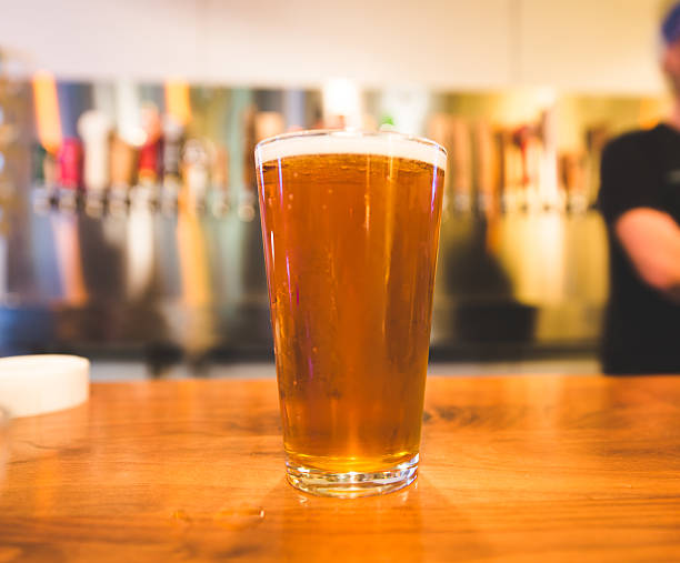 A freshly-poured pint of beer sits on the bar at a tap house or bar. Taps and bartender in background.