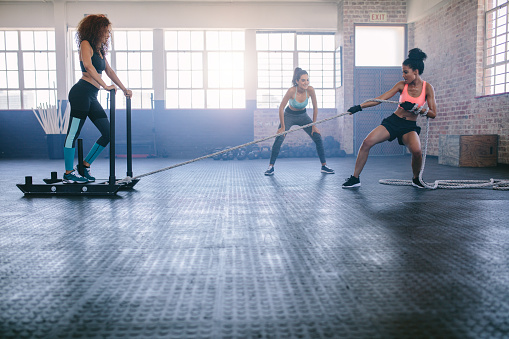 Shot of woman standing on sled with other pulling in gym. Three young females doing physical training at healthclub.