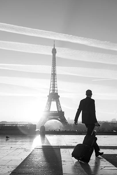 A beautiful sunrise takes place in Paris behind the iconic Eiffel Tower and a young businessman walks with his business luggage and contemplates the iconic monument. Concept of worldwide business travel, mobility and business confidence. In Black and White mode.