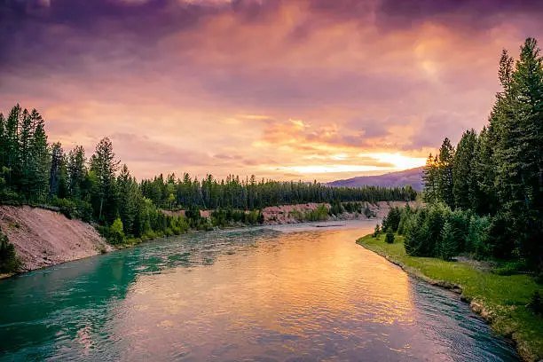 This is a horizontal, color photograph of the scenic landscape of Glacier National Park at sunset. The Middle Fork Flathead River reflects the vibrant colors in the sky. The river banks are full of green conifer trees. Photographed in spring with a Nikon D800 DSLR camera.