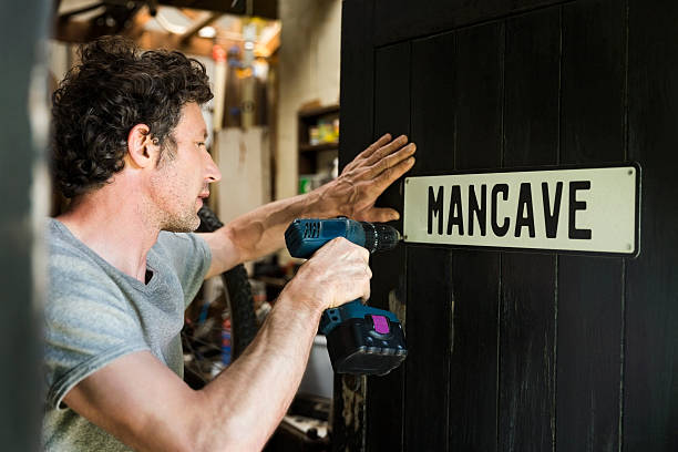 Mature man fixing mancave sign on wooden door Mature man fixing mancave sign on wooden door. Male repairman using power tool. He is working at workshop. door repairman men manual worker stock pictures, royalty-free photos & images