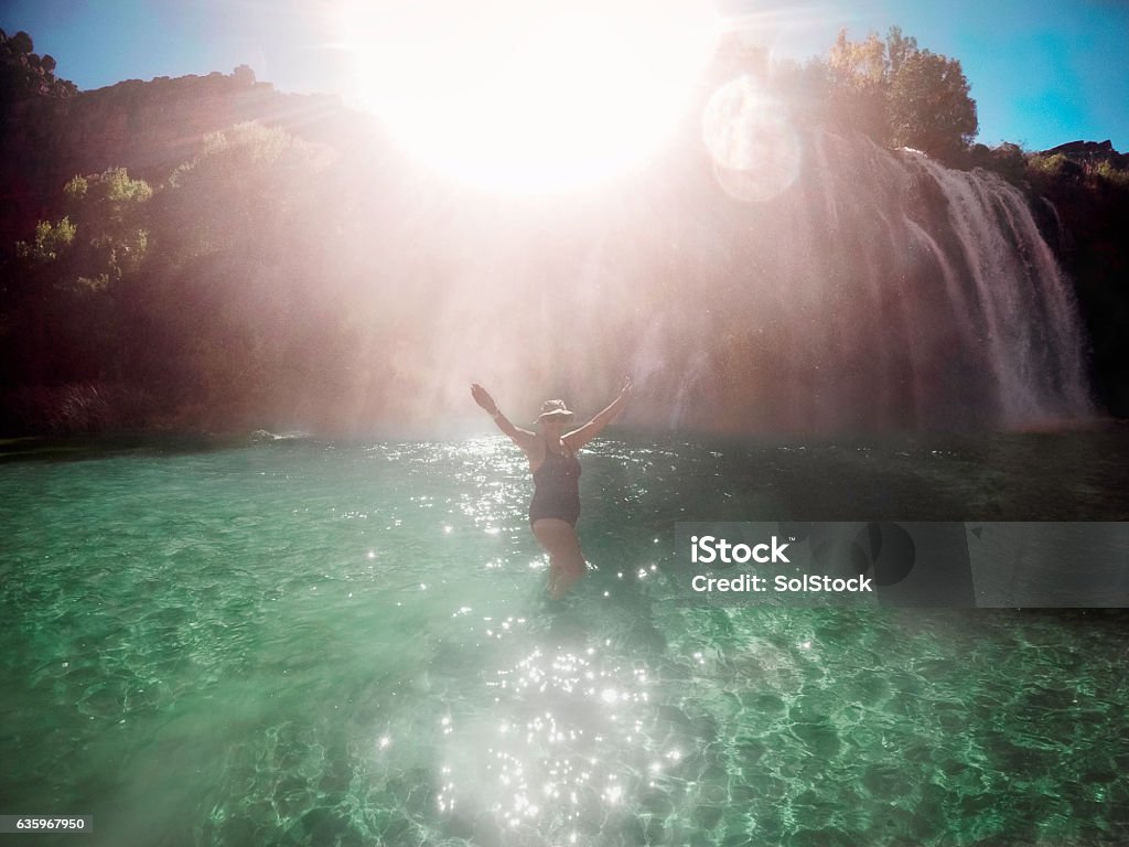 Keeping Cool in the Grand Canyon Senior woman standing in a pool of water from a water fall in the Grand Canyon. Sun shines from over the of a waterfall creating a bright light effect 50-54 Years Stock Photo
