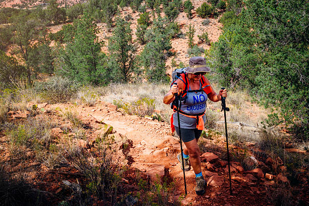 Trekking Her Way to the Top Senior woman hiking up a footpath using walking poles in the Grand Canyon. Trees and red rocky terrain can be seen below. The woman is wearing a sun hat red rocks state park arizona photos stock pictures, royalty-free photos & images