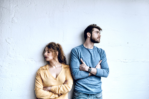 serious young couple looking in opposite directions, leaning on rustic white wall in loft