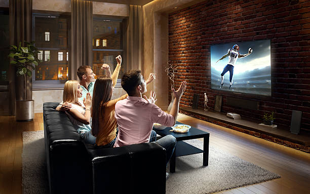 Couples watching American football game at home :biggrin:Two couples are cheering while watching American football game at home. They are sitting on a sofa in the modern living room faced to a big TV set on the front wall. excited football fan cheering for his team on tv stock pictures, royalty-free photos & images