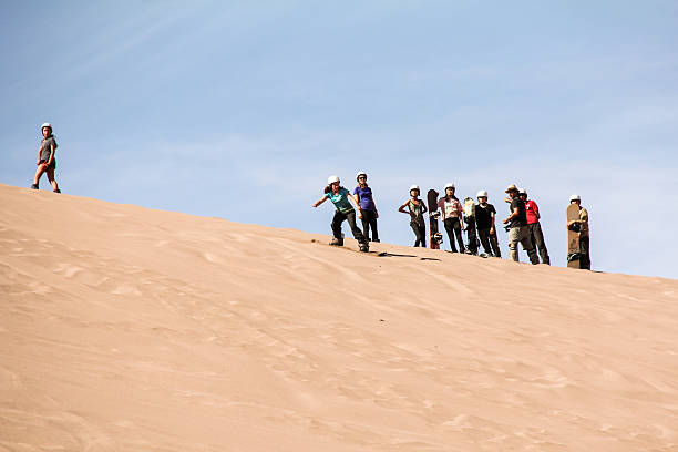 A person stands barefoot on the crest of a wind-sculpted sand dune, carrying a long wooden sandboard across their shoulders. They face sweeping, sunlit dunes that roll into the distance under a clear deep-blue sky, with footprints and soft shadows tracing the sandy ridges. The scene evokes solitude, adventure and the vast scale of the desert landscape.