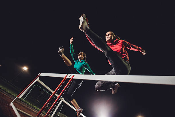 Hurdling Young Athletes Two young female athletes doing hurdle race training on a running track at dusk. hurdling track event stock pictures, royalty-free photos & images