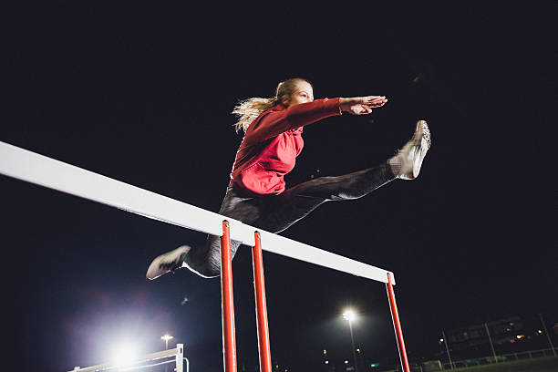 Hurdling Young Athlete Young female athlete doing hurdle race training on a running track at dusk. womens track stock pictures, royalty-free photos & images