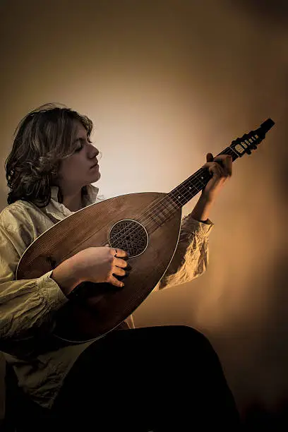 Young man with long blond hair sitting in dark environment and playing an Old Oud, Guitar Lute and looking at it. He is dressed in white rustic baroque shirt with ruffle edging to collars.