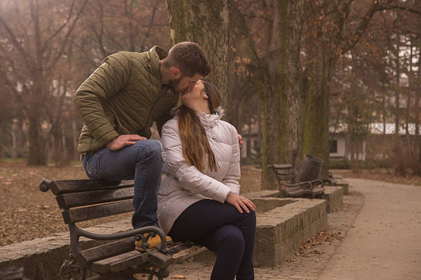 young adult couple sitting in park bench kissing. outdoors day winter
