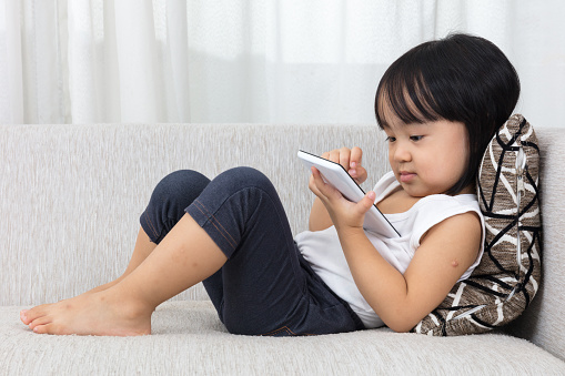 Asian Chinese little girl lying on the sofa with mobile phone in the living room at home.