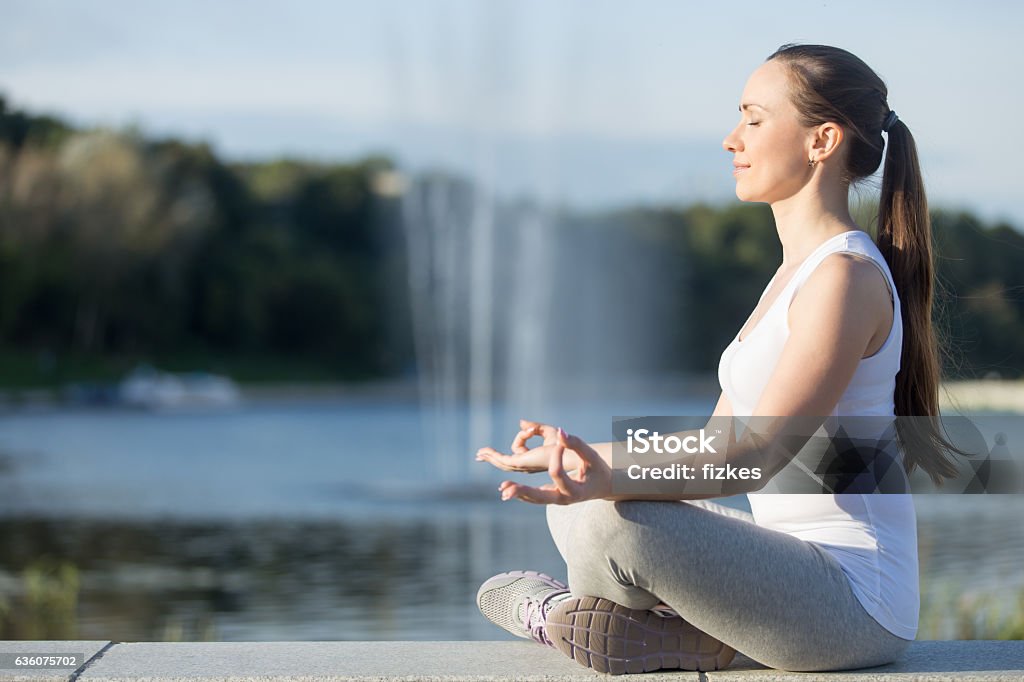Easy Seat against fountain Profile portrait of sporty beautiful young woman practicing yoga, sitting in Easy Seat exercise, Sukhasana pose, working out, wearing sportswear, outdoor full length, fountain background. Copy space Activity Stock Photo
