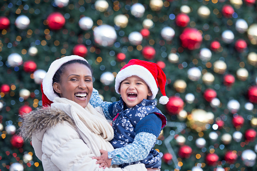 Young mother carrying her little child on hands, standing against Christmas decorated tree. Woman and her daughter wearing red Santa hats and looking over the camera with a smile.