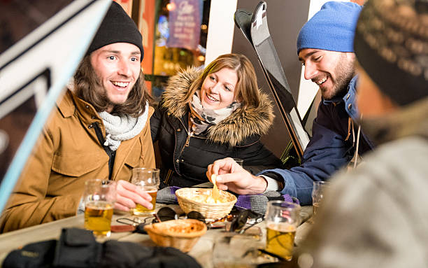 Happy friends drinking beer having fun at ski resort chalet Happy friends drinking beer and eating chips - Cheerful people having fun at bar restaurant by skiing resort with snow equipment - Friendship concept on warm night filter with focus on young woman apres ski stock pictures, royalty-free photos & images