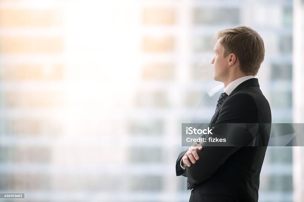 Confident businessman waiting for the meeting Successful businessman in suit standing in office with hands crossed on chest, looking through window at big city buildings, planning new projects, waiting for meeting to start. Copy space for text Businessman Stock Photo