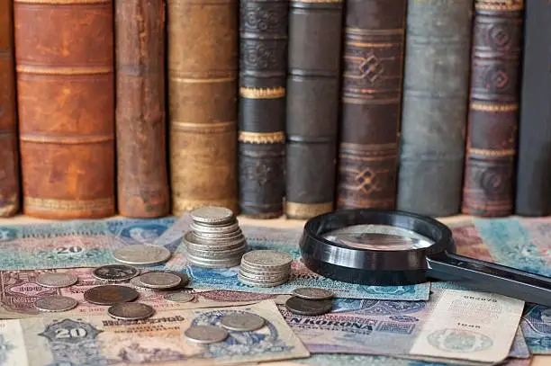 Old coins and magnifying glass are lying on banknotes with antiquarian books on the background.