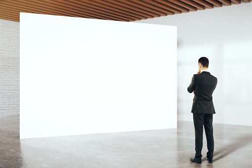 Thoughtful businessman in suit looking at blank billboard in white birck interior. Mock up, 3D Rendering