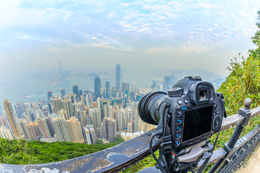 Close up of a professional camera on the tripod while photographing the Victoria Harbour from The Victoria Peak in Hong Kong. Fisheye lens with focus on the camera and background skyline blurred.