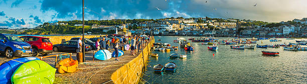St Ives, UK - August 5, 2016: Crowds of holiday makers, day trippers and tourists enjoying the summer sunshine on the harbour wall of St. Ives, Cornwall. Composite panoramic image created from eleven contemporaneous sequential photographs. 