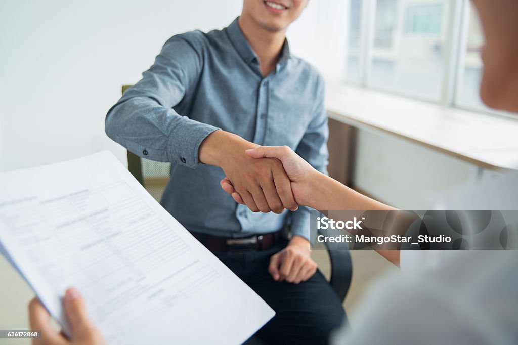 Smiling Asian businessman shaking partners hand Smiling Asian businessman wearing shirt sitting in office and shaking hand of female partner. Woman holding document Agreement Stock Photo