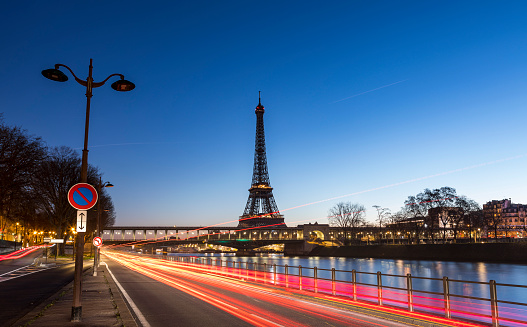 An early morning sunrise takes place behind the majestic Eiffel Tower in the city of Paris in France. Situated near the Seine river, the Eiffel Tower is one of the most important monument in the World. The long exposure creates red tail lights coming from the cars on the move.