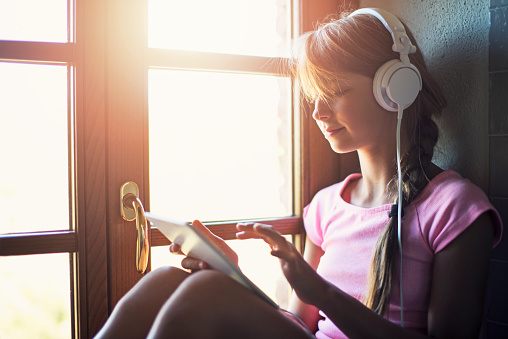 Smiling teenage girl using tablet next to a window. The girl is wearing white headphones.