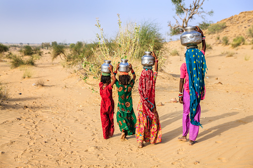 Indian little girls crossing sand dunes and carrying on their heads water from local well, Thar Desert, Rajasthan, India. Rajasthani women and children often walk long distances through the desert to bring back jugs of water that they carry on their heads. 