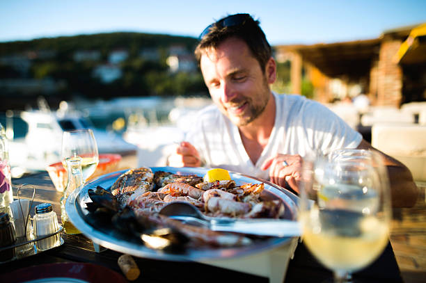homme en vacances, assis dehors au coucher du soleil, manger des fruits de mer - wine food fish seafood photos et images de collection