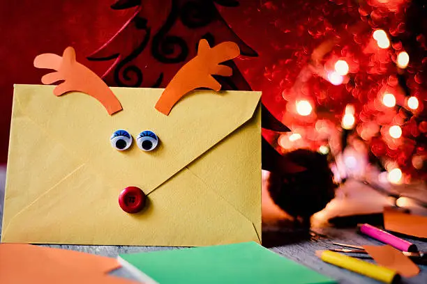 closeup of an envelope customized as a reindeer face with a button as nose and paperboard antlers, containing a letter to santa, placed next to a wooden christmas tree