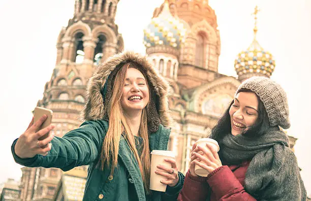 Happy girlfriends taking winter selfie at " Savior on Spilled Blood " church in Saint Petersburg - Friendship concept with girls having fun together drinking coffee outdoor - Focus on left young woman