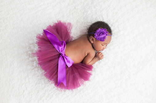 Portrait of a one month old, sleeping, newborn, baby girl. She is wearing a purple tutu and sleeping on a white blanket.