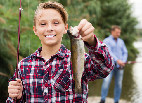 Cheerful teenage boy holding catch freshwater fish in hands on weekend