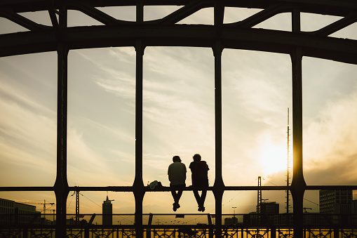 Two men sit on a steel bridge looking into the sunset.