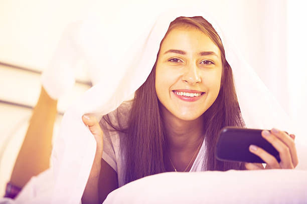 улыбающаяся девушка с смартфон, скрывающихся в постель - directly below looking women human head стоковые фото и изображения
