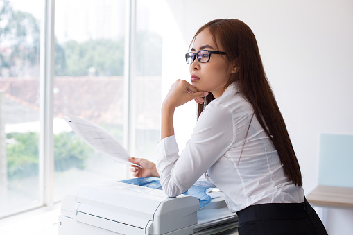 Closeup of thoughtful Asian business woman leaning on photocopier, holding document and looking nowhere with office window and blurred city view outside. Side view.