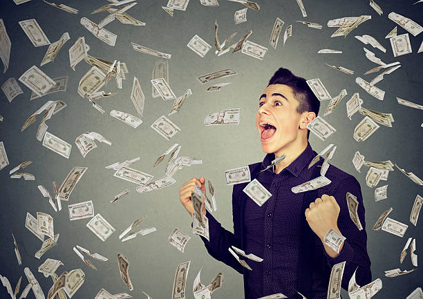 Man celebrates success under money rain falling down dollar banknotes Portrait happy man exults pumping fists ecstatic celebrates success under money rain falling down dollar banknotes isolated on gray wall background jackpot photos stock pictures, royalty-free photos & images