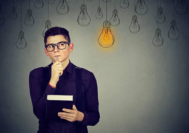 Handsome thinking man in glasses looking up with light idea bulb above head isolated on gray wall background.