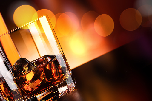 Glass and bottle of whisky on the bar, with ice cubes in glass, on dark background. Blurred yellow and red lights in background. Reflection on the surface. Horizontal orientation. Close-up.