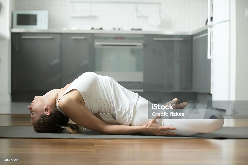 Fish yoga pose Sporty young attractive woman practicing yoga, lying in Fish exercise, Matsyasana pose, working out, wearing white sportswear, indoor full length, home interior background Active Lifestyle Stock Photo