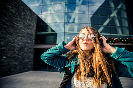 Young woman with sunglasses holding headphones an listening to music in front of a bulding made of glass
