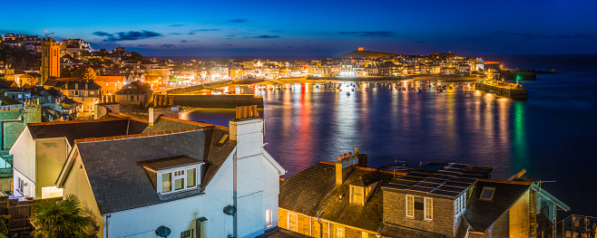 Panoramic dusk view across the rooftops and church towers of St. Ives to the warm glow of the quayside shops, pubs and restaurants around the famous harbour.