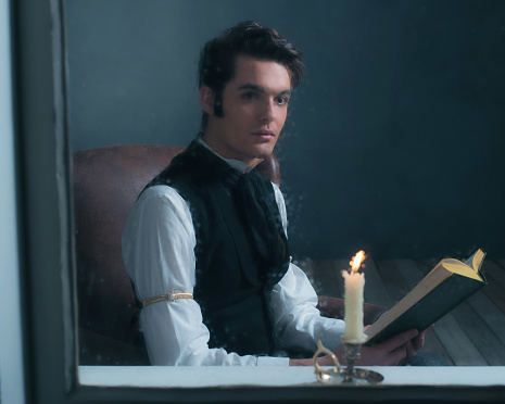 Vintage victorian man sitting by rainy window reading book with candlelight.