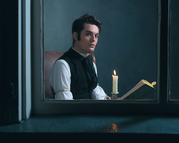 Retro victorian man reading book by candlelight sitting by window.
