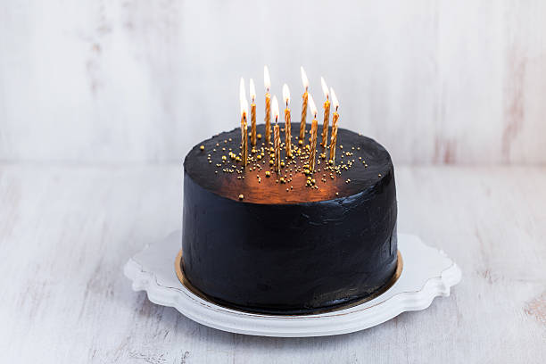 istock Black round birthday cake with burning golden candles 636401052