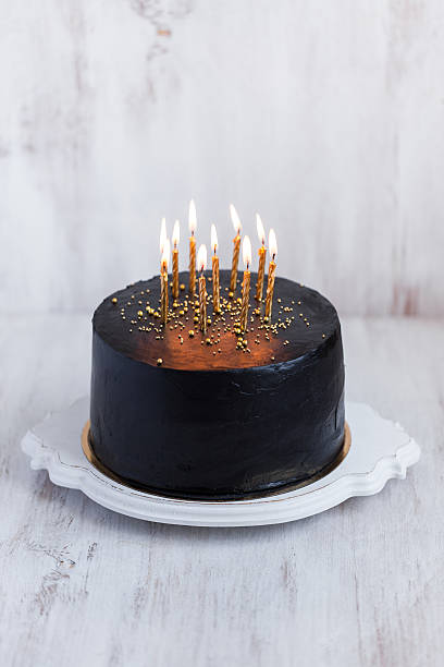 istock Black round birthday cake with burning golden candles 636401094