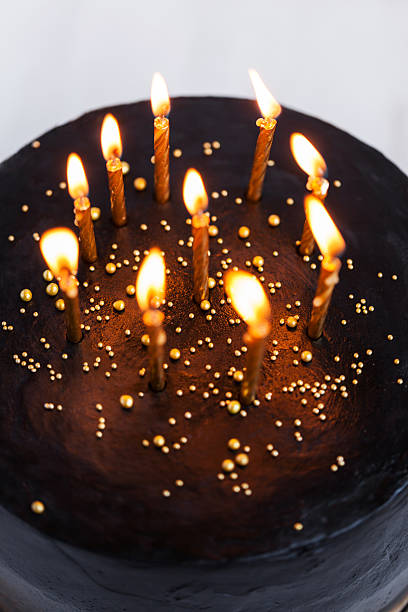 istock Black round birthday cake with burning golden candles 636401196