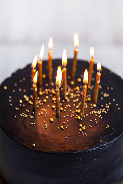 istock Black round birthday cake with burning golden candles 636401676